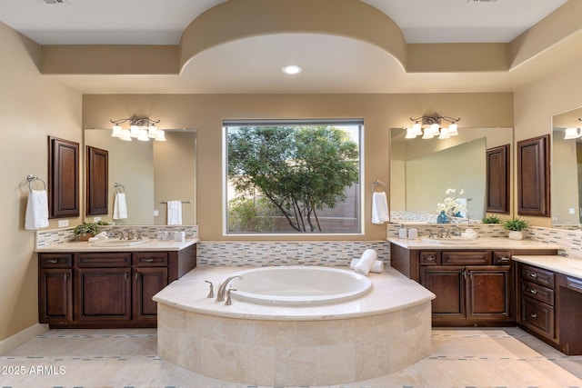 bathroom with two vanities, a garden tub, backsplash, and a sink