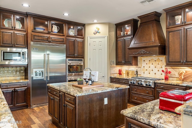 kitchen with built in appliances, custom range hood, visible vents, dark brown cabinets, and dark wood finished floors