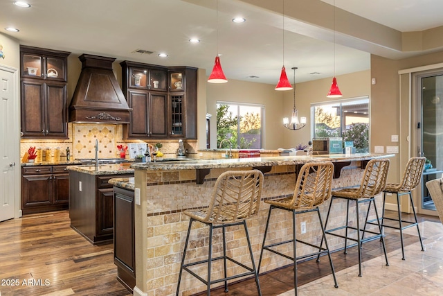 kitchen with tasteful backsplash, light stone counters, premium range hood, visible vents, and a center island with sink
