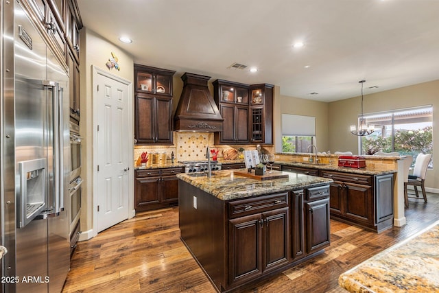 kitchen featuring tasteful backsplash, appliances with stainless steel finishes, visible vents, premium range hood, and a center island with sink