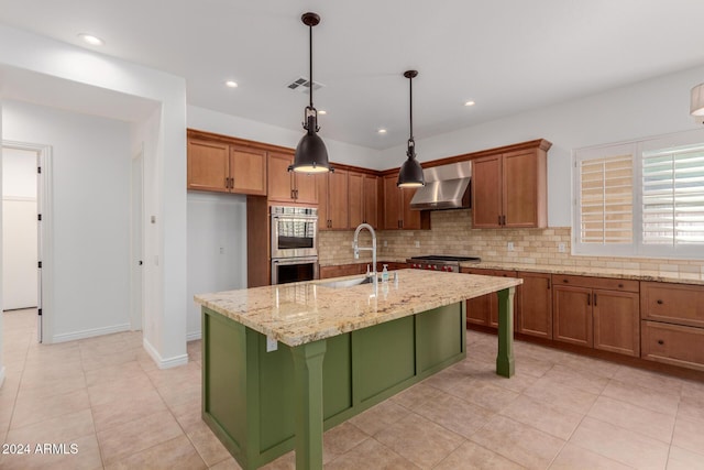 kitchen featuring light stone countertops, sink, wall chimney range hood, double oven, and a kitchen island with sink