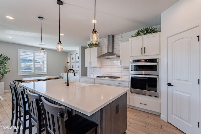 kitchen with white cabinetry, a kitchen island with sink, stainless steel appliances, decorative backsplash, and wall chimney exhaust hood