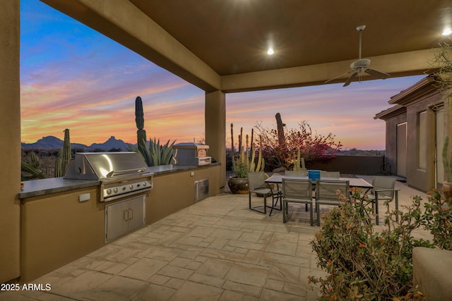 patio terrace at dusk with area for grilling, a mountain view, ceiling fan, and an outdoor kitchen