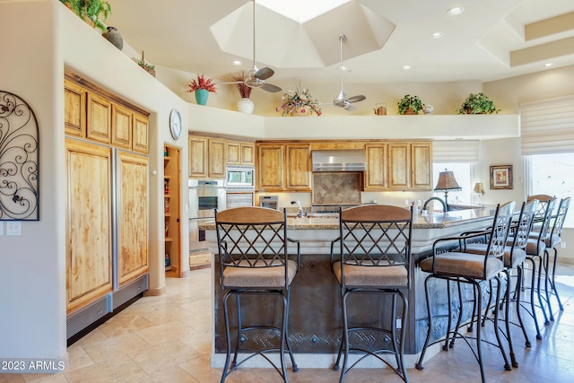 kitchen featuring light stone countertops, appliances with stainless steel finishes, a towering ceiling, wall chimney exhaust hood, and ceiling fan