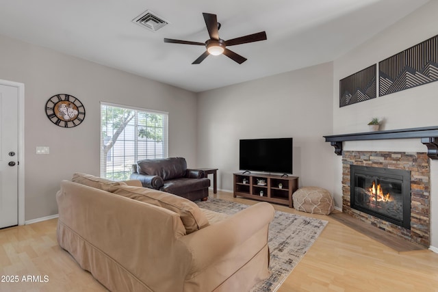 living room with visible vents, light wood-style floors, baseboards, a stone fireplace, and a ceiling fan