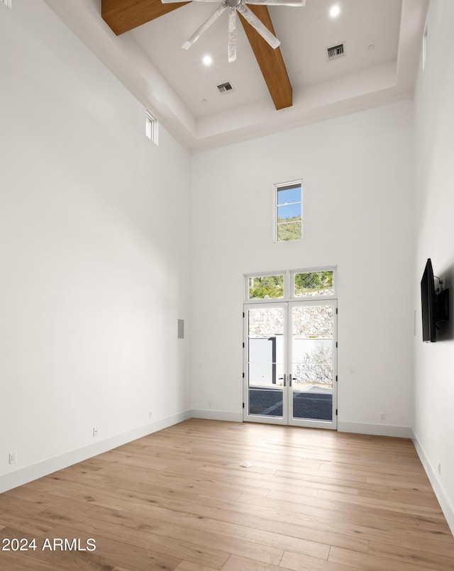 unfurnished living room with beamed ceiling, a high ceiling, ceiling fan, light wood-type flooring, and french doors