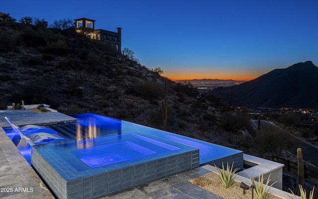 pool at dusk with an in ground hot tub, a mountain view, and a patio area