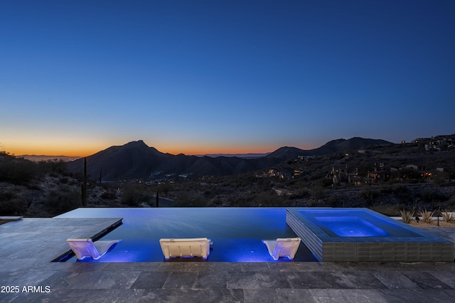 pool at dusk featuring a mountain view, a patio area, and an in ground hot tub