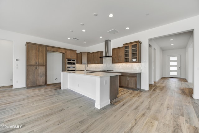 kitchen with wall chimney range hood, a center island with sink, and light wood-type flooring