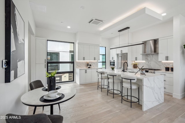 kitchen featuring sink, white cabinets, hanging light fixtures, a kitchen island with sink, and wall chimney exhaust hood