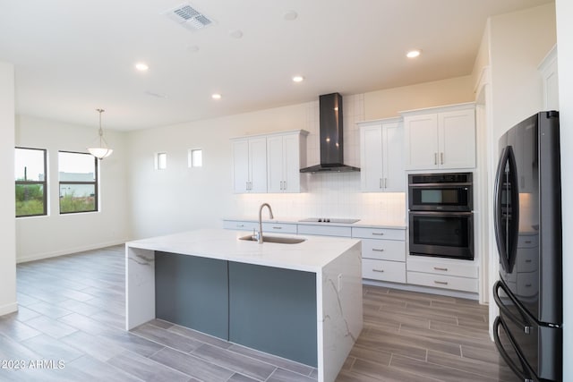 kitchen featuring white cabinetry, wall chimney range hood, light stone counters, fridge, and a center island with sink