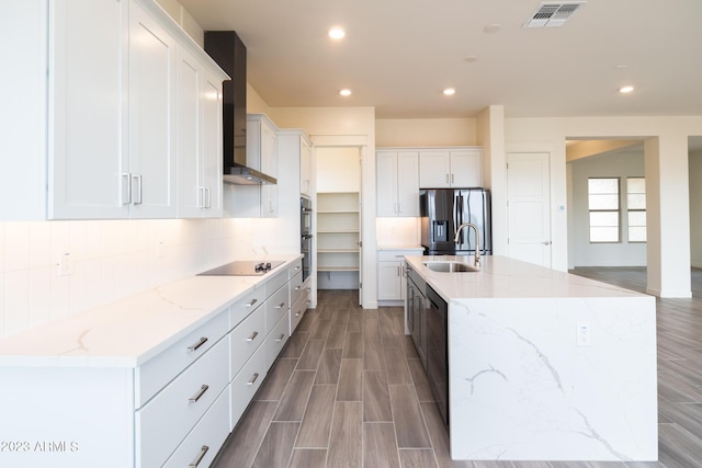 kitchen with a large island with sink, black appliances, wall chimney range hood, sink, and white cabinetry