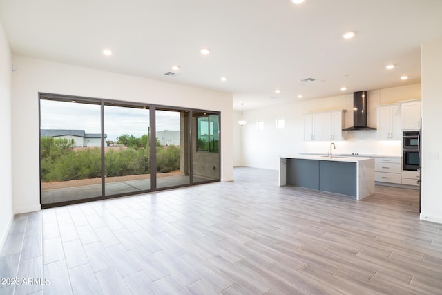 kitchen featuring wall chimney exhaust hood, double oven, sink, a center island with sink, and white cabinets