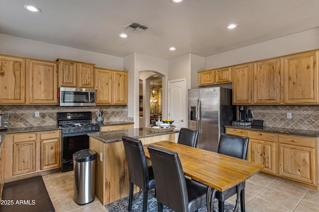 kitchen with light tile patterned floors, stainless steel appliances, a kitchen island, decorative backsplash, and dark stone counters