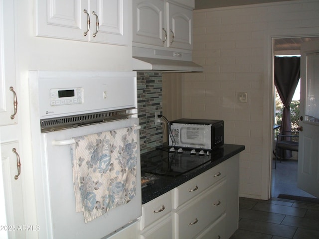 kitchen featuring decorative backsplash, dark countertops, white oven, under cabinet range hood, and white cabinetry