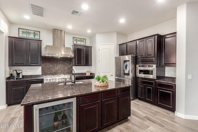 kitchen featuring an island with sink, sink, wine cooler, stainless steel appliances, and wall chimney range hood