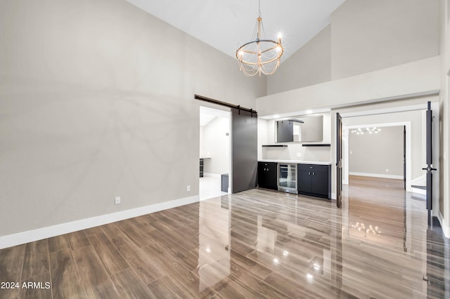 unfurnished living room with a barn door, wine cooler, high vaulted ceiling, a chandelier, and hardwood / wood-style flooring