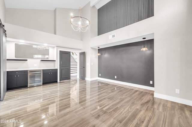 unfurnished living room with beverage cooler, a barn door, light hardwood / wood-style flooring, high vaulted ceiling, and a notable chandelier