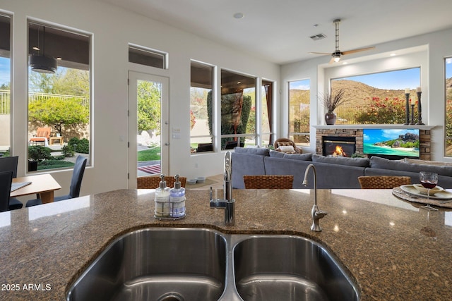 kitchen featuring visible vents, open floor plan, a stone fireplace, dark stone countertops, and a ceiling fan