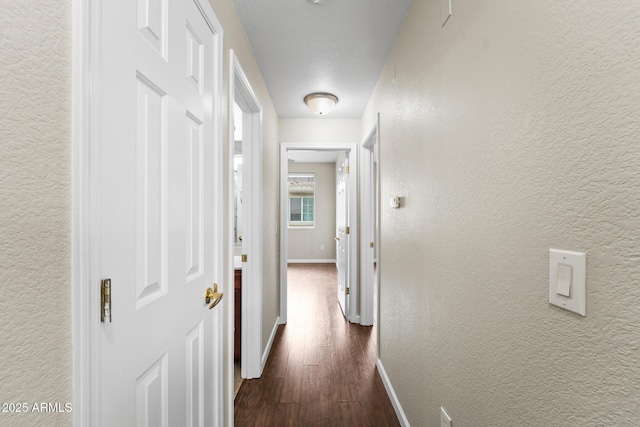 corridor featuring a textured wall, dark wood-style flooring, and baseboards