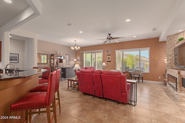 living room featuring light tile patterned flooring, ceiling fan with notable chandelier, a stone fireplace, and sink