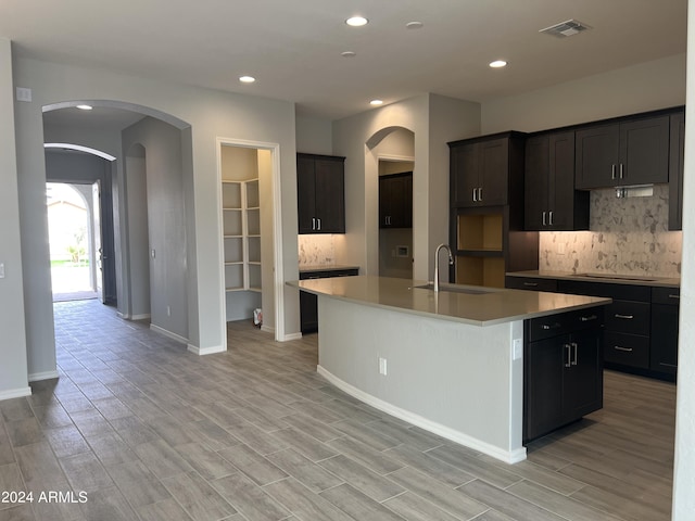 kitchen with arched walkways, light countertops, decorative backsplash, a kitchen island with sink, and a sink