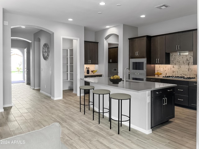 kitchen featuring stainless steel gas stovetop, a center island with sink, sink, tasteful backsplash, and dark brown cabinets