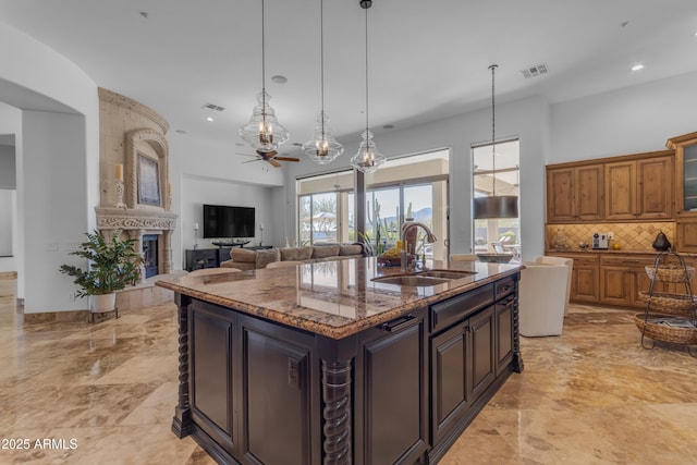 kitchen with light stone counters, a sink, an island with sink, a tiled fireplace, and glass insert cabinets