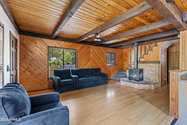 living room with wooden walls, light hardwood / wood-style flooring, a wood stove, ceiling fan, and beam ceiling