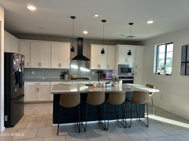 kitchen featuring appliances with stainless steel finishes, white cabinetry, a kitchen island with sink, and wall chimney range hood