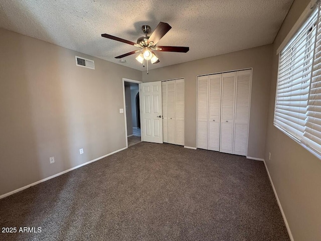 unfurnished bedroom with dark colored carpet, two closets, ceiling fan, and a textured ceiling