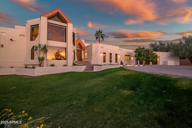 back house at dusk featuring a balcony, a patio, and a lawn