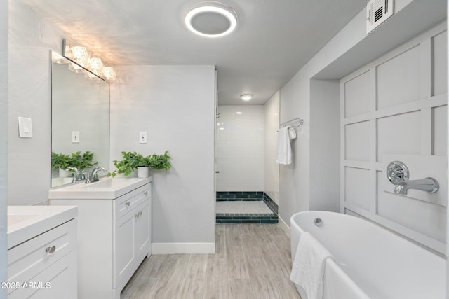 bathroom with wood finished floors, two vanities, tiled shower, baseboards, and a soaking tub