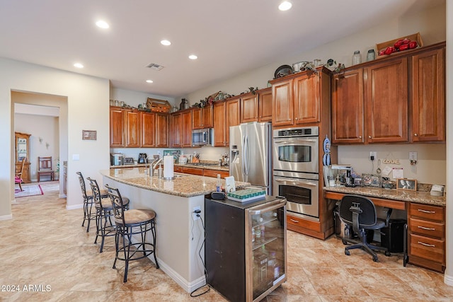 kitchen with a kitchen bar, light stone countertops, stainless steel appliances, beverage cooler, and an island with sink