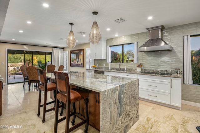 kitchen with white cabinets, a kitchen island, light stone countertops, pendant lighting, and wall chimney range hood