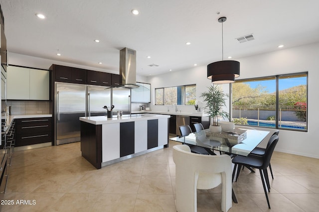 kitchen featuring hanging light fixtures, appliances with stainless steel finishes, island exhaust hood, a kitchen island with sink, and decorative backsplash