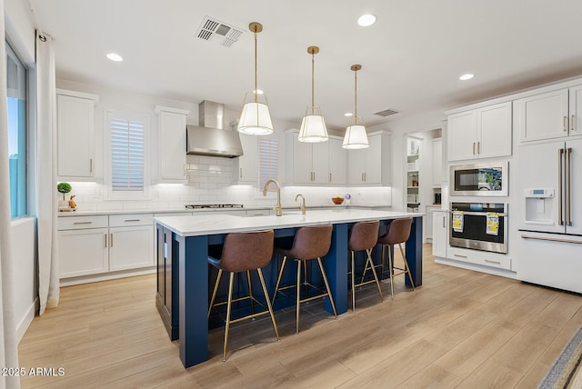 kitchen featuring built in microwave, wall chimney range hood, oven, an island with sink, and white cabinets