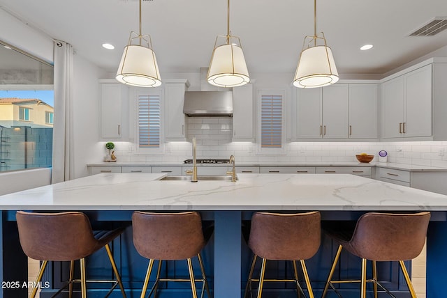 kitchen featuring wall chimney exhaust hood, a kitchen island with sink, white cabinets, and hanging light fixtures