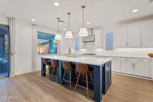 kitchen featuring a kitchen bar, wall chimney range hood, pendant lighting, white cabinetry, and an island with sink