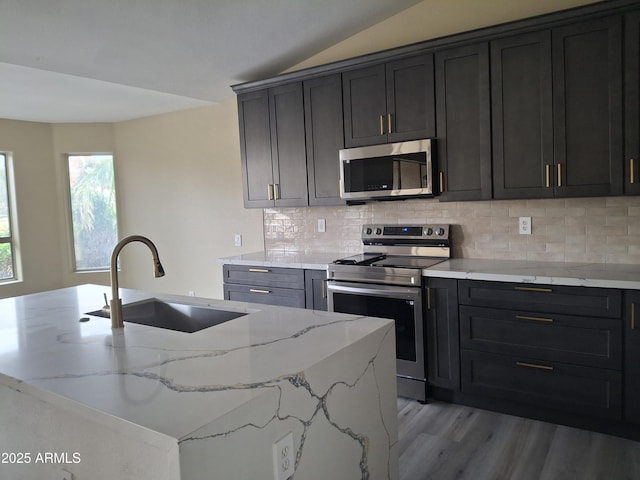 kitchen with lofted ceiling, sink, appliances with stainless steel finishes, a kitchen island with sink, and light stone counters