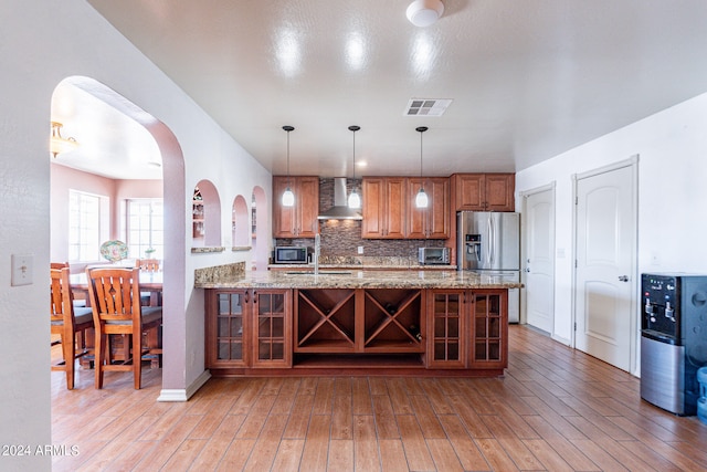 kitchen featuring light stone countertops, appliances with stainless steel finishes, light hardwood / wood-style flooring, hanging light fixtures, and wall chimney range hood