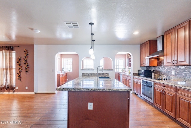 kitchen featuring appliances with stainless steel finishes, wall chimney range hood, a kitchen island with sink, and a wealth of natural light