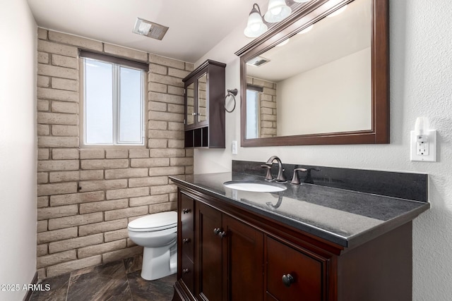 bathroom with visible vents, toilet, vanity, and brick wall