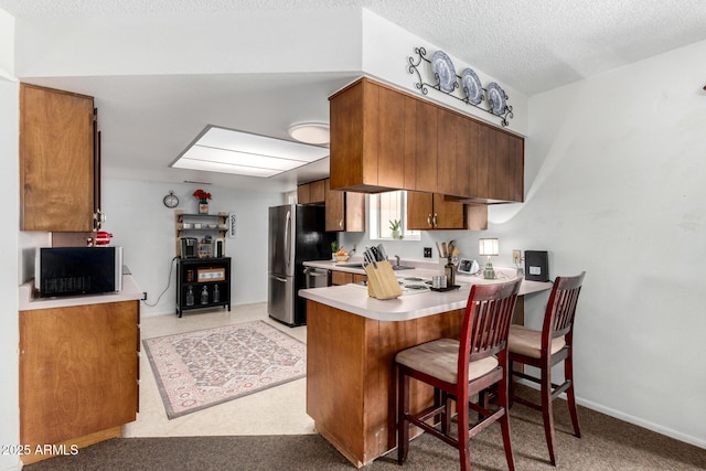 kitchen with a textured ceiling, freestanding refrigerator, a peninsula, brown cabinetry, and light countertops