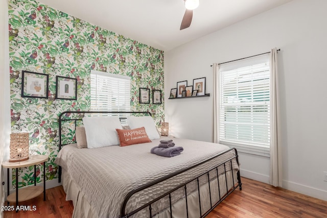 bedroom with a ceiling fan, wood finished floors, wallpapered walls, baseboards, and an accent wall