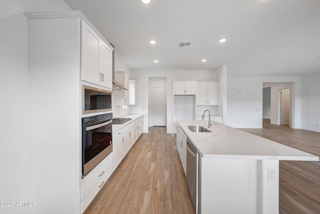 kitchen with light wood-style flooring, a kitchen island with sink, a sink, appliances with stainless steel finishes, and decorative backsplash