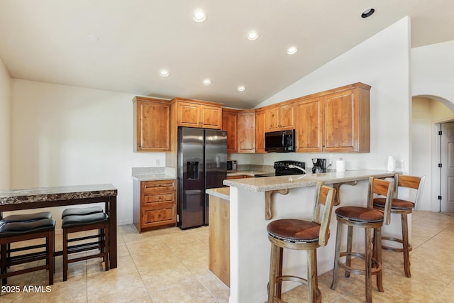 kitchen with arched walkways, light stone counters, brown cabinets, black appliances, and a kitchen bar