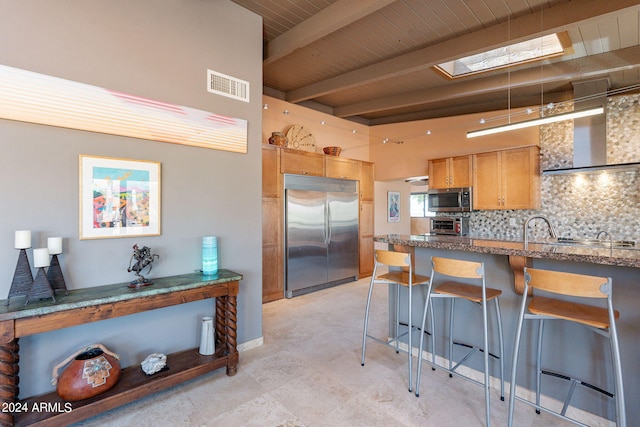 kitchen featuring appliances with stainless steel finishes, a breakfast bar, beamed ceiling, a skylight, and wall chimney range hood