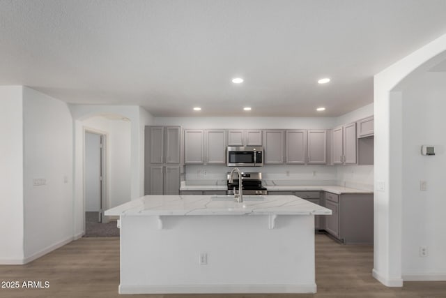 kitchen featuring arched walkways, light stone counters, gray cabinetry, a sink, and appliances with stainless steel finishes