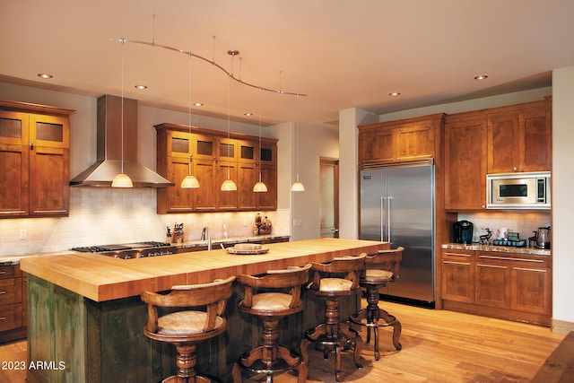 kitchen featuring built in appliances, butcher block counters, a breakfast bar area, and wall chimney range hood